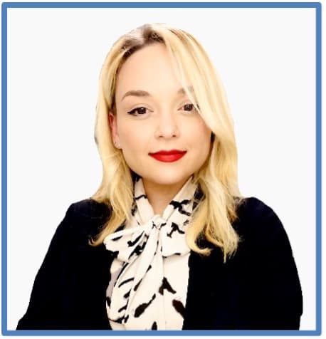 Blonde woman with red lipstick wearing a black blazer and a patterned bow blouse.